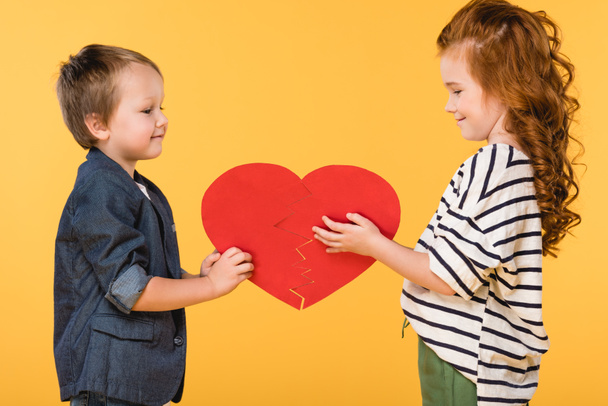 side view of smiling kids holding red paper heart together isolated on yellow, st valentines day concept - Zdjęcie, obraz