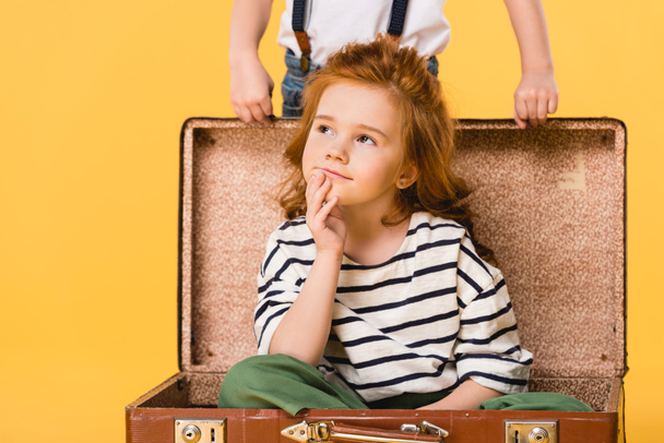 partial view of dreamy kid sitting in suitcase with boy standing near by isolated on yellow - Photo, image