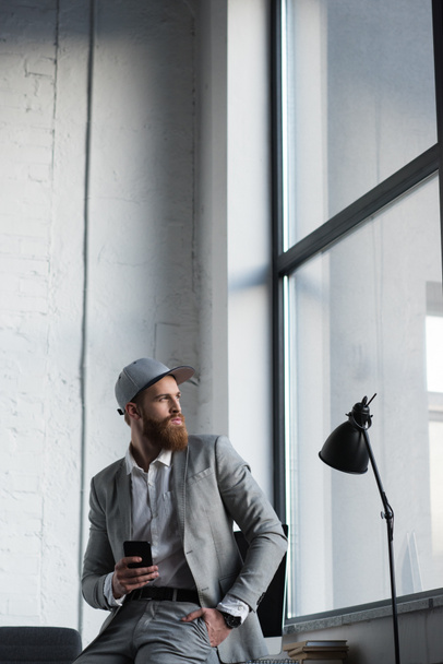 businessman in baseball cap holding smartphone and looking away - Valokuva, kuva