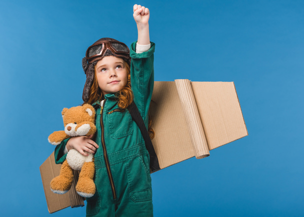 retrato de niño lindo en traje piloto con oso de peluche y alas de avión de papel hechas a mano aisladas en azul
 - Foto, Imagen