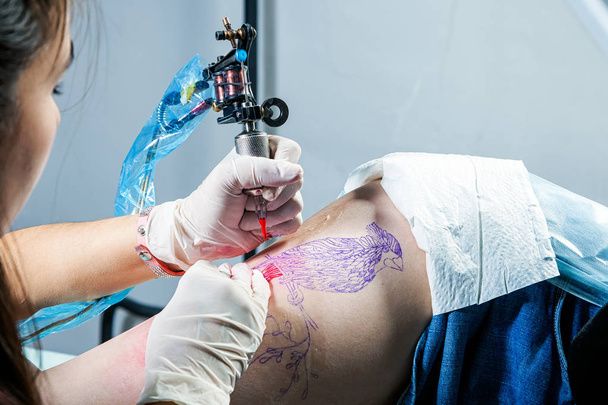 Primer plano del tatuaje maestro dibuja la pintura roja en el tatuaje de los clientes. Tatuaje artista sosteniendo una máquina de tatuaje de metal en guantes estériles y trabajando en la estera azul profesional
. - Foto, Imagen
