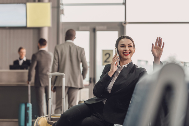 young businesswoman waiting for plane at airport lobby while talking by phone and waving hand - Foto, imagen