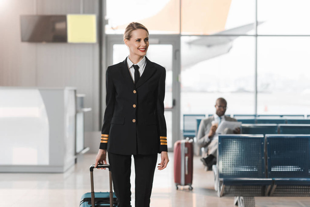улыбающаяся женщина-пилот с чемоданом, идущая мимо вестибюля аэропорта
 - Фото, изображение