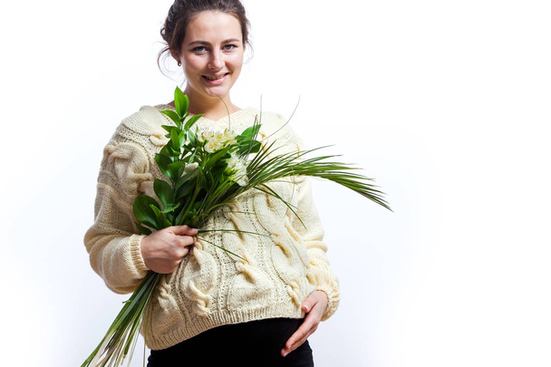 Young beautiful pregnant woman in a black dress and a knitted sweater of milky color from a natural wool holds a bouquet of flowers in her hands and is weakened on an isolated white background, side view - Photo, Image