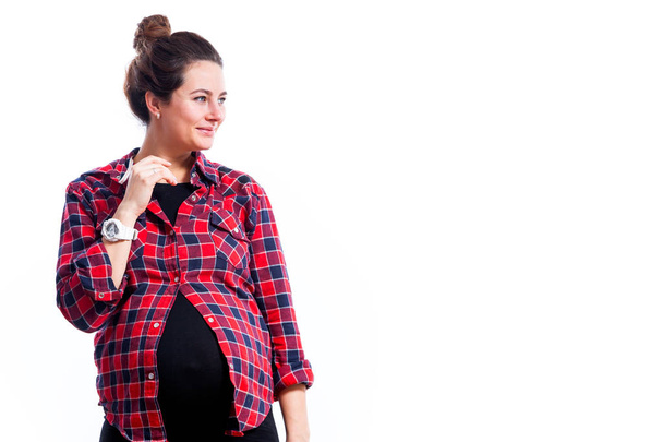 Young dark-haired pregnant woman in black dress and plaid shirt smiling and posing on isolated white background - Photo, Image