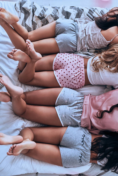 top view of four women in pajamas lying on bed, partial view of human legs and butts in shorts - Foto, afbeelding