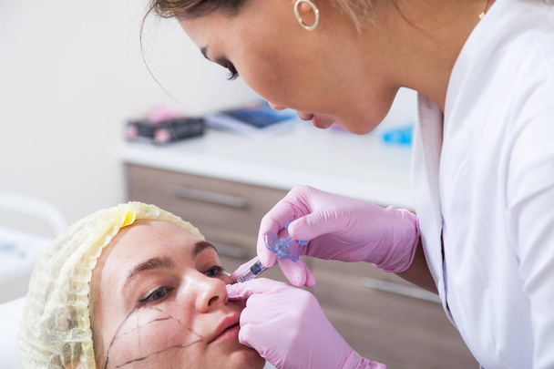 Közeli kép egy sötét hajú nő egy ázsiai kozmetikus fecskendővel, Botox injekció intravénás az arccsontja, egy fiatal nő az orvosi rendelőben alakjának beállításához - Fotó, kép