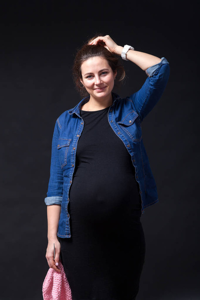 Dark-haired mooie jongedame op de laatste zwangerschap in spijkerbroek shirt en zwarte jurk glimlacht en bezit in handen niet-afhankelijke roze trui met breinaalden op een zwarte achtergrond van de geïsoleerde - Foto, afbeelding