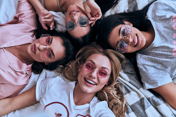 Top view of beautiful young women smiling while lying on bed in pajamas and sun glasses - Foto, afbeelding