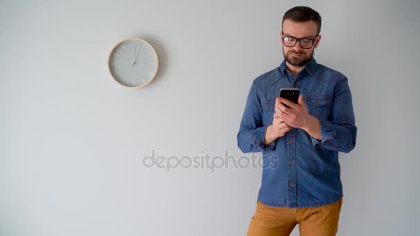 Brodaty mężczyzna przy użyciu smartfona pod ścianą, szary z zegarem - Materiał filmowy, wideo