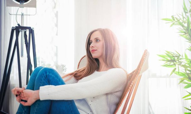 Woman at home sitting on modern chair near window relaxing in living room - Photo, Image