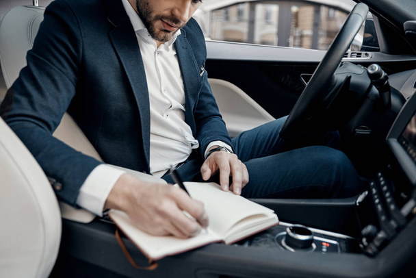 Close up of young man in full suit writing notes in personal organizer while sitting inside of the car - Foto, Bild