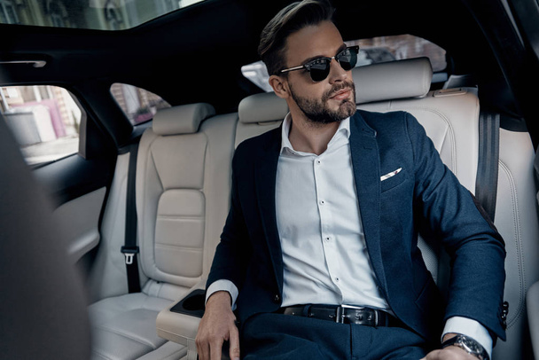 Handsome young man in suit and glasses looking away while sitting in car - Foto, afbeelding