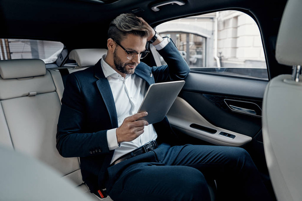 confident young businessman in suit using digital tablet while sitting in car - Foto, immagini