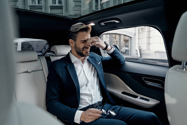 Frustrated young man in formal wear massaging nose and keeping eyes closed while sitting in the car - Fotoğraf, Görsel