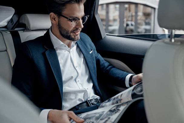 Handsome young businessman in suit reading a newspaper while sitting in car - Photo, Image