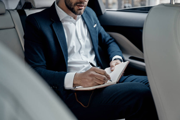 Close up of young man in formal wear writing notes in diary while sitting in car - Valokuva, kuva