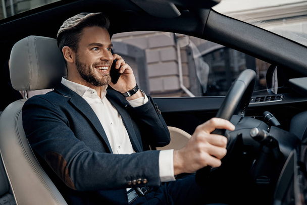 Handsome young man in formal wear talking on his smart phone and smiling while driving a luxury car - Foto, Bild