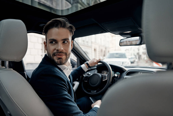 Handsome young man in suit looking over shoulder and smiling while driving a car - Fotoğraf, Görsel