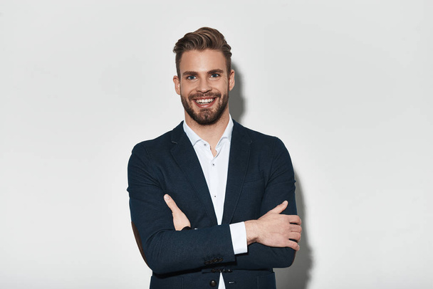 Good looking young man in full suit keeping arms crossed and looking at camera with smile while standing against grey background - Fotoğraf, Görsel