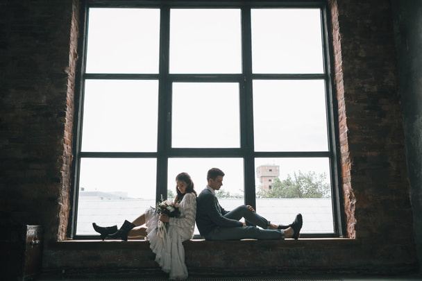 La mariée et le marié sont assis dos à dos sur le rebord de la fenêtre. Joyeux nouveaux mariés. Œuvre d'art - Photo, image