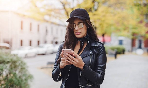 Elegáns lány visel a fekete kupakot és a bőrdzseki SMS gépelés mobiltelefon kültéri - Fotó, kép