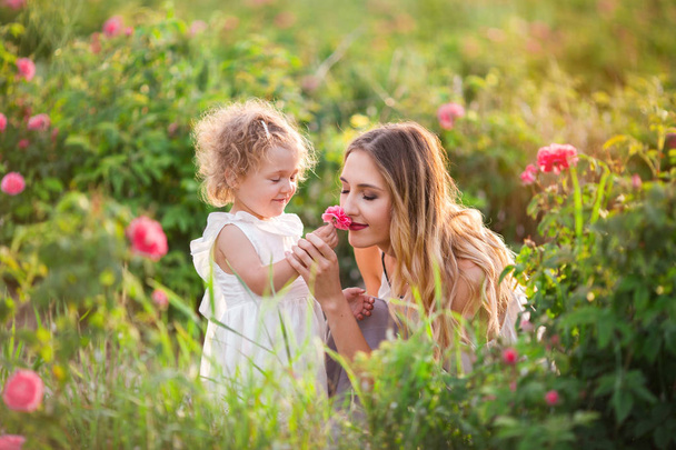 Beautiful child girl with pretty young mother are walking in spring garden with pink blossom roses flowers - Photo, Image