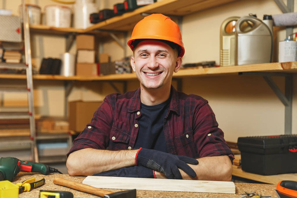 Handsome smiling caucasian young man in plaid shirt, black T-shirt, orange protective helmet, gloves working in carpentry workshop at wooden table place with piece of wood, hammer, different tools. - Photo, Image