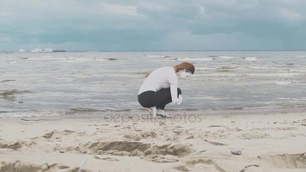 Kumlu deniz sahilinde beyaz boya kaplı sanatsal kadın çömelmiş - Video, Çekim