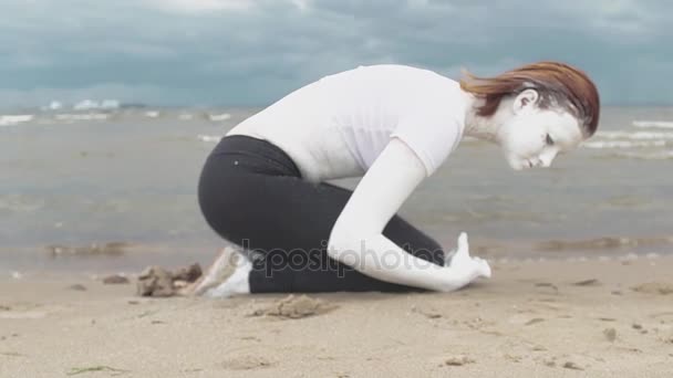 Artista de performance cubierto de pintura blanca agachado en la orilla del mar de arena
 - Metraje, vídeo