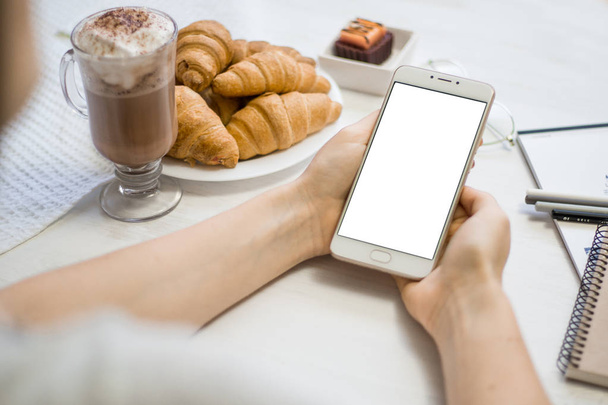 Attrappe eines Smartphones in Mädchenhänden in einem Café auf dem Hintergrund eines hölzernen weißen Tisches Nahaufnahmetelefon mit weißem Bildschirm - Foto, Bild