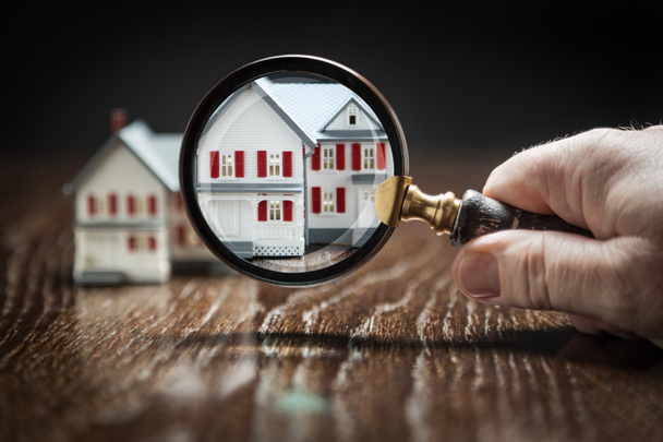 Hand Holding Magnifying Glass Up To Model Home on Reflective Wooden Surface. - Photo, Image