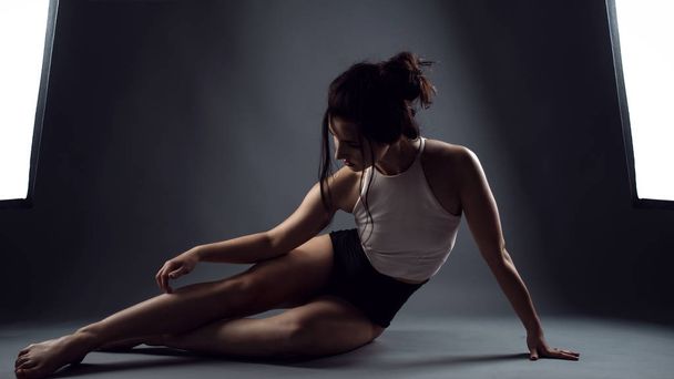 Retrato de una joven atleta en bragas negras de cintura alta y parte superior blanca sentada en el suelo y tocándose la pierna
 - Foto, Imagen