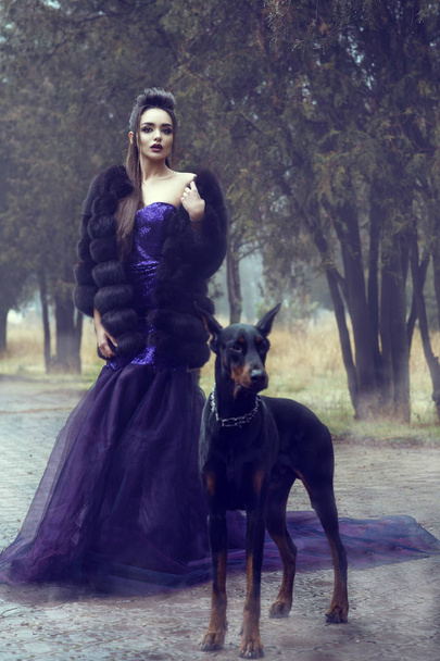 Young glam beautiful lady wearing luxurious sequin violet evening gown and fur coat standing on the alley in the park with her Doberman pinscher dog in front of her - Photo, Image