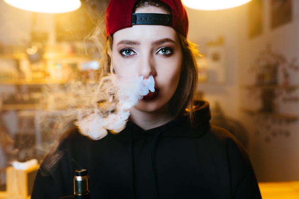 Young pretty woman in red cap smoke an electronic cigarette at the vape shop. Hip-hop style. Closeup. - Photo, Image