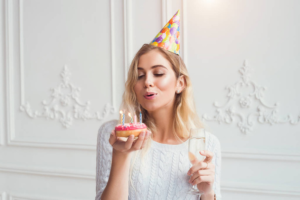 帽子を被った女の子が誕生日ドーナツを保持しています。女性はシャンパンを飲むキャンドルで blowind です。誕生日ケーキ。パーティーのお祝い。休日の気分. - 写真・画像