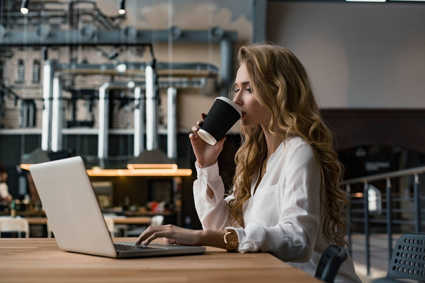 Μια γυναίκα εργάζεται για το laptop της. Ψάχνει για πληροφορίες στο διαδίκτυο. Η γυναίκα πίνει καφέ. Μια εργάσιμη ημέρα. - Φωτογραφία, εικόνα