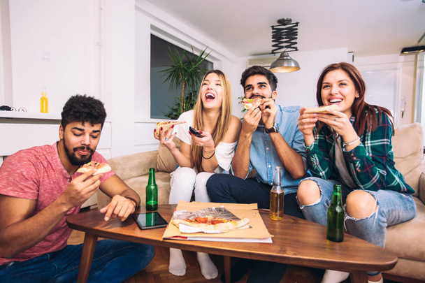 Φίλοι και πίτσα. Τέσσερις νέοι χαρούμενα τρώγοντας πίτσα στο σπίτι. - Φωτογραφία, εικόνα