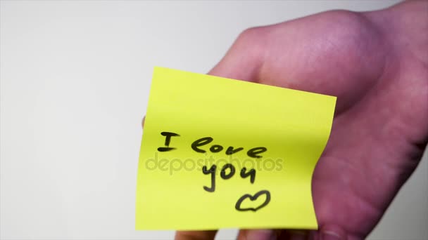 Note with I love you. Blank yellow sticky note with I love you sign pined on a cork bulletin board. Inscription on the sticker I love you on white background. Mans hand put blank yellow sticker - Video