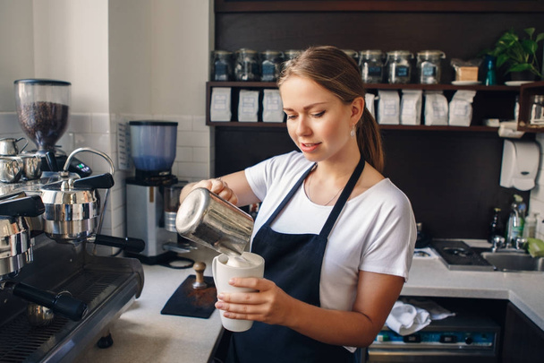Πορτρέτο της χαμογελαστός νεαρός καυκάσιος γυναίκα barista στην ποδιά χύνοντας γάλα στον καφέ. Ιδιοκτήτης της επιχειρηματικής ιδέας.  - Φωτογραφία, εικόνα