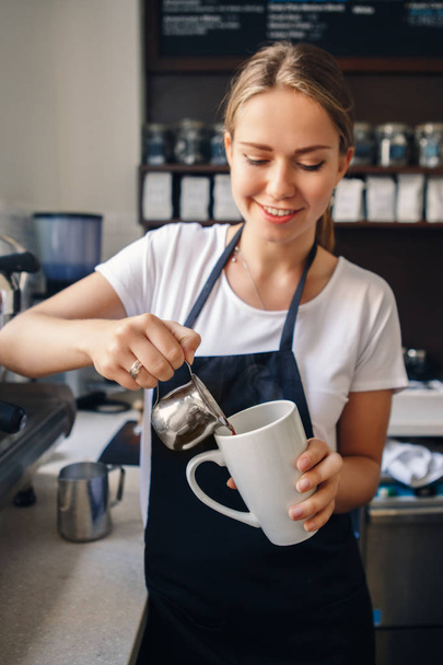 Portret uśmiechnięta młoda kaukaski kobieta barista w fartuch nalewanie kawy w kubek. Koncepcja biznesowa właściciela.  - Zdjęcie, obraz
