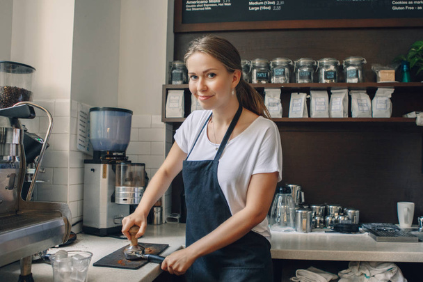 Young Caucasian woman barista using tamper to press ground coffee into portafilter to make espresso hot drink. Small local business work in cafe. - Photo, Image