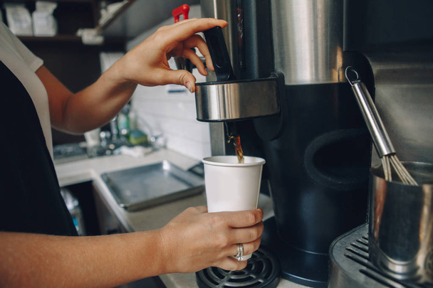 Young Caucasian barista hands holding paper cup making coffee using coffee machine. Woman pouring coffee from professional espresso machine. Small business and person at work concept - Photo, Image
