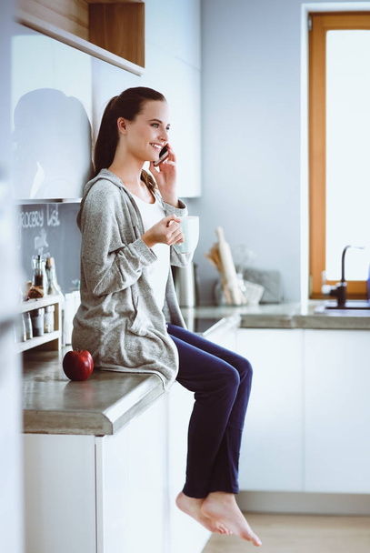 Woman using mobile phone sitting in modern kitchen. - Foto, afbeelding
