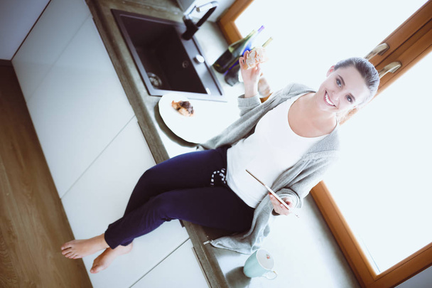 Beautiful young woman using a digital tablet in the kitchen. - 写真・画像