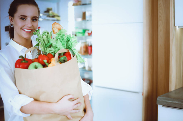 野菜と食料品の買い物袋を持っている若い女性.キッチンに立って.台所の女性がカメラを見て - 写真・画像