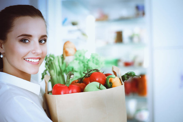 Młoda kobieta trzyma torbę na zakupy z warzywami.Stojąc w kuchni. Kobieta w kuchni patrząca w kamerę - Zdjęcie, obraz