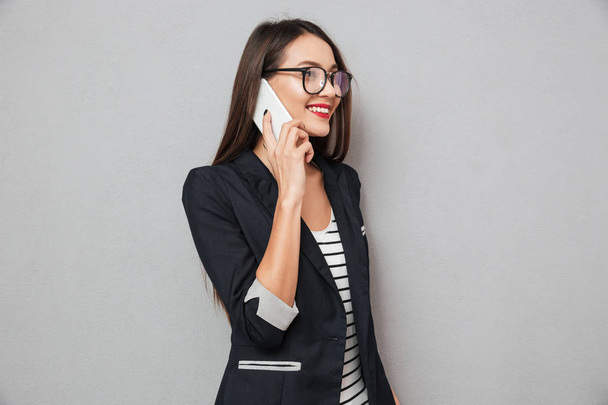 Smiling asian business woman in eyeglasses talking by smartphone - Фото, изображение