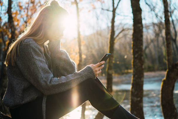 Zijaanzicht van zonovergoten vrouw surfen smartphone in vallen bos - Foto, afbeelding