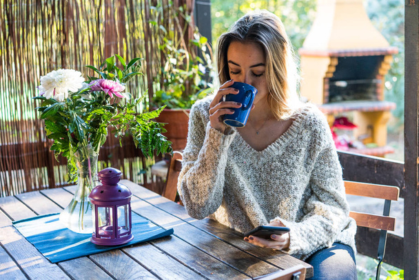 自宅でスマート フォンを見ながらコーヒーを飲むセーターの若い女性 - 写真・画像